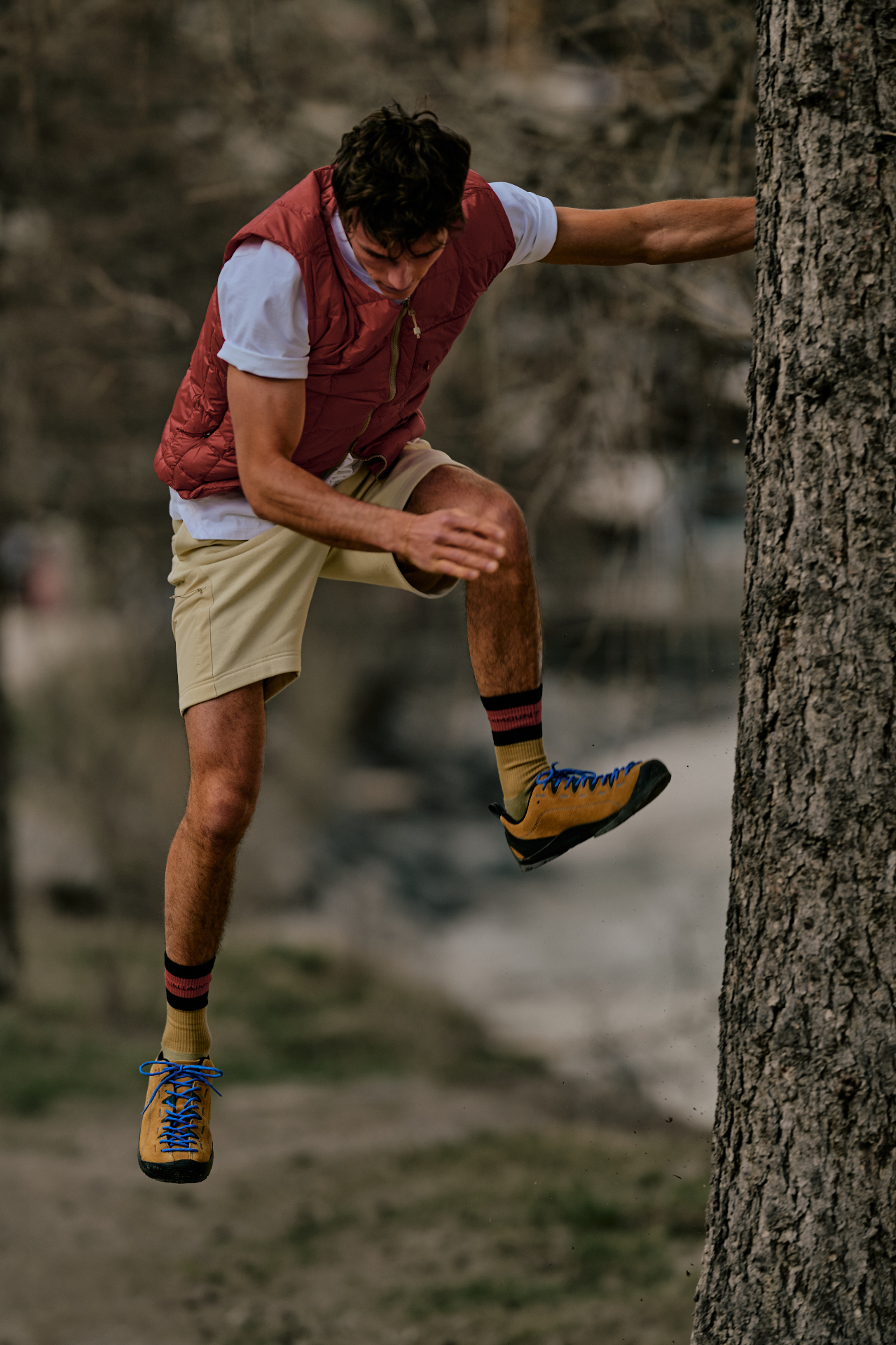 Man in burgundy quilted gilet, white tee and approach shoes mid-jump near a tree