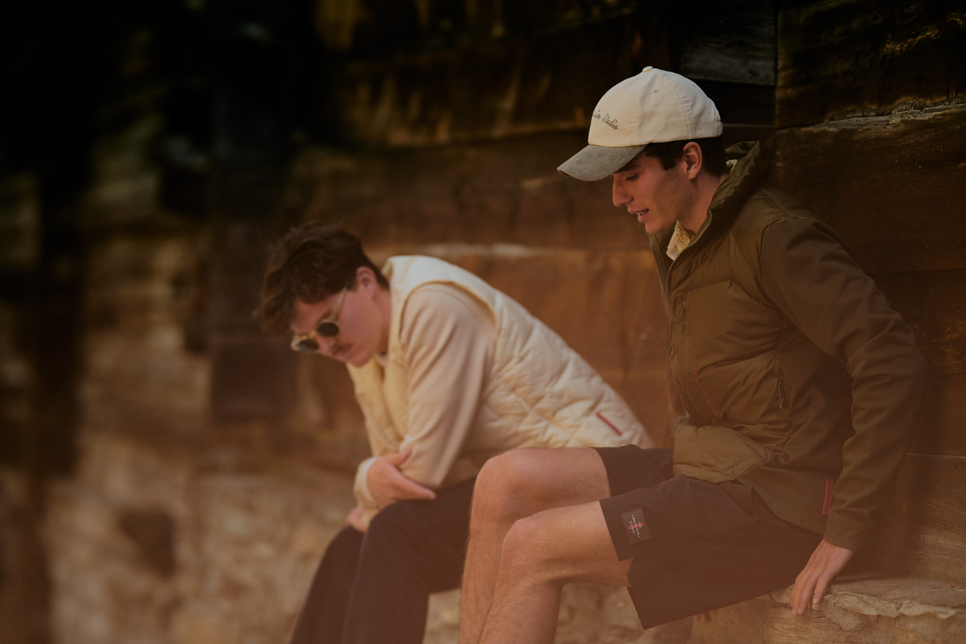 Two men seated on a Zermatt chalet step in cream quilted vest and olive puffer with corduroy cap